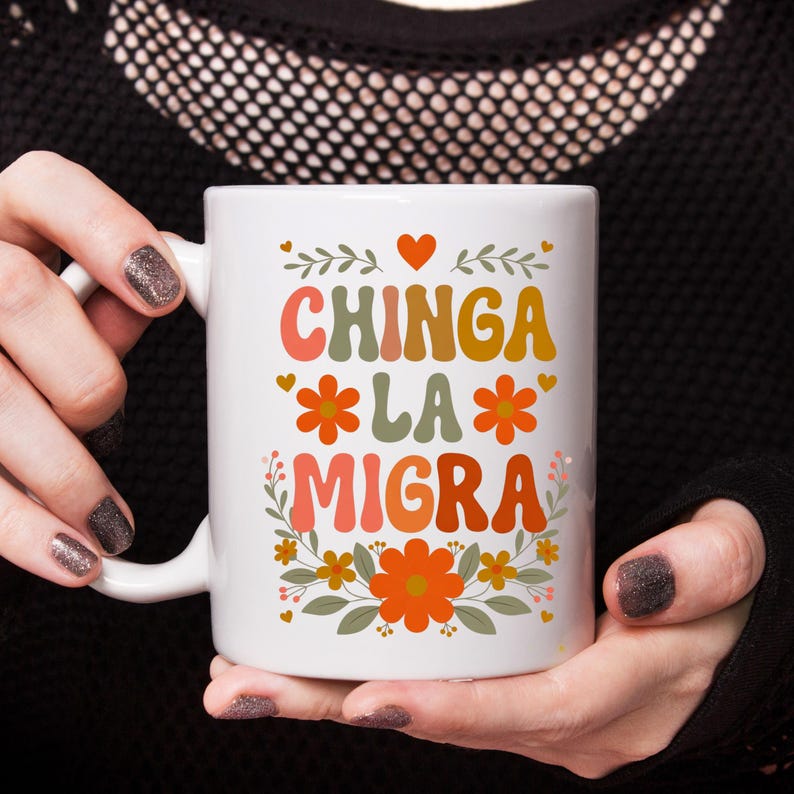May include: White ceramic mug with the text "CHINGA LA MIGRA" in colorful, retro-style lettering. The design includes floral accents and hearts. The mug is held by a person with dark nail polish.