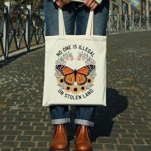 May include: Cream-colored tote bag featuring a monarch butterfly design with sunflowers and the text "NO ONE IS ILLEGAL ON STOLEN LAND." The butterfly is orange and black, surrounded by yellow sunflowers and pink flowers.