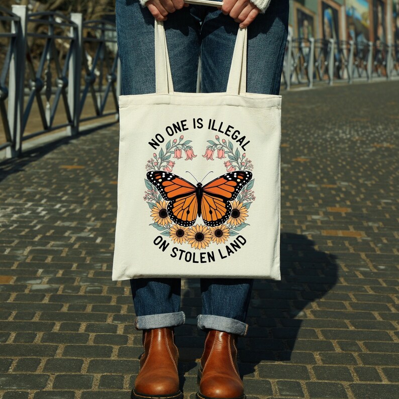 May include: Cream-colored tote bag featuring a monarch butterfly design with sunflowers and the text "NO ONE IS ILLEGAL ON STOLEN LAND." The butterfly is orange and black, surrounded by yellow sunflowers and pink flowers.