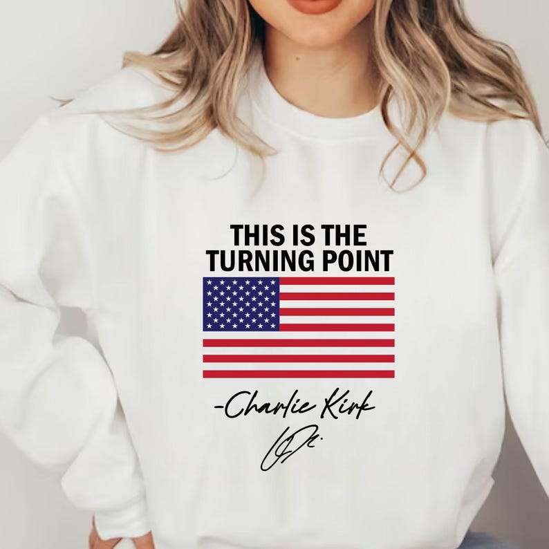May include: White sweatshirt featuring the text "THIS IS THE TURNING POINT" and an American flag design. The signature "-Charlie Kirk" is printed below the flag.