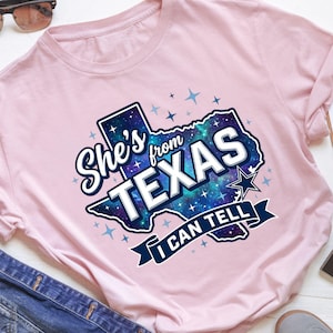 Pode incluir: Uma t-shirt rosa claro com um gr&aacute;fico do estado do Texas preenchido com um padr&atilde;o de gal&aacute;xia azul e roxo. O texto "She's from TEXAS I CAN TELL" est&aacute; em letras brancas e azul-escuras, rodeado por pequenos &iacute;cones de estrelas azul-claras.