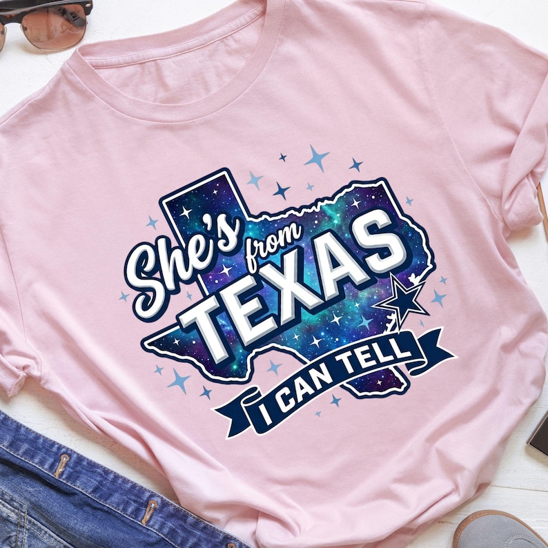 Pode incluir: Uma t-shirt rosa claro com um gr&aacute;fico do estado do Texas preenchido com um padr&atilde;o de gal&aacute;xia azul e roxo. O texto "She's from TEXAS I CAN TELL" est&aacute; em letras brancas e azul-escuras, rodeado por pequenos &iacute;cones de estrelas azul-claras.