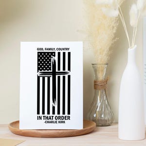 May include: A white rectangular print with a black and white design. The design features a cross over an American flag. The text at the top reads "GOD, FAMILY, COUNTRY" and the text at the bottom reads "IN THAT ORDER -CHARLIE KIRK".
