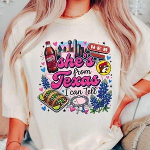 Pode incluir: Uma t-shirt cor de creme com um gr&aacute;fico que inclui o texto 'she's from Texas I can Tell'. O design apresenta uma garrafa de Dr Pepper, um horizonte urbano, um sinal H-E-B, um log&oacute;tipo Buc-ee's, tacos, tremo&ccedil;os e uma pulseira com berloques do Texas.