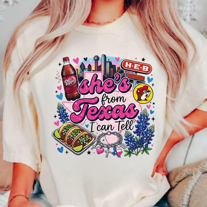 Pode incluir: Uma t-shirt cor de creme com um gr&aacute;fico que inclui o texto 'she's from Texas I can Tell'. O design apresenta uma garrafa de Dr Pepper, um horizonte urbano, um sinal H-E-B, um log&oacute;tipo Buc-ee's, tacos, tremo&ccedil;os e uma pulseira com berloques do Texas.