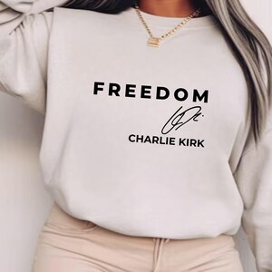 May include: A cream-colored sweatshirt featuring the word "FREEDOM" in black, bold lettering. Beneath the word is a signature and the name "CHARLIE KIRK" in black. The garment is a neutral tone.