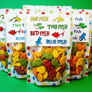 May include: Several clear cellophane bags filled with colorful fish-shaped crackers. Each bag has a white label with the text "One Fish, Two Fish, Red Fish, Blue Fish" and illustrations of fish. The bags are on a green surface.