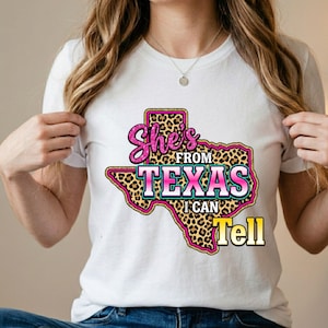 Pode incluir: Uma t-shirt branca de manga curta com um gr&aacute;fico do estado do Texas com preenchimento de estampado de leopardo. O texto "She's FROM TEXAS I CAN Tell" est&aacute; escrito em letras cor-de-rosa e brancas com efeito brilhante, centrado na frente.