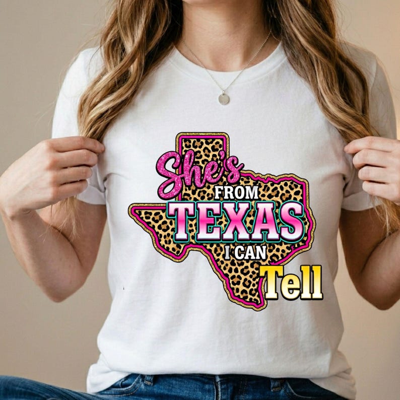 Pode incluir: Uma t-shirt branca de manga curta com um gr&aacute;fico do estado do Texas com preenchimento de estampado de leopardo. O texto "She's FROM TEXAS I CAN Tell" est&aacute; escrito em letras cor-de-rosa e brancas com efeito brilhante, centrado na frente.