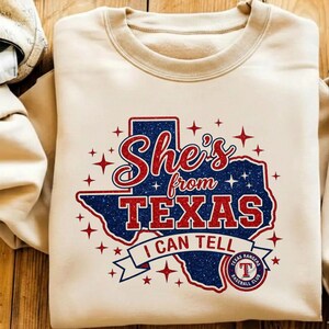 Pode incluir: Uma camisola bege de gola redonda com um gr&aacute;fico do estado do Texas em glitter azul. O texto "She's from TEXAS I CAN TELL" est&aacute; em glitter vermelho, acompanhado por estrelas vermelhas brilhantes e o log&oacute;tipo do Texas Rangers Baseball Club.