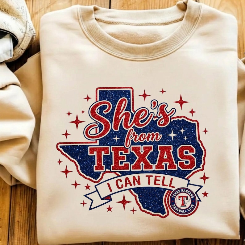 Pode incluir: Uma camisola bege de gola redonda com um gr&aacute;fico do estado do Texas em glitter azul. O texto "She's from TEXAS I CAN TELL" est&aacute; em glitter vermelho, acompanhado por estrelas vermelhas brilhantes e o log&oacute;tipo do Texas Rangers Baseball Club.