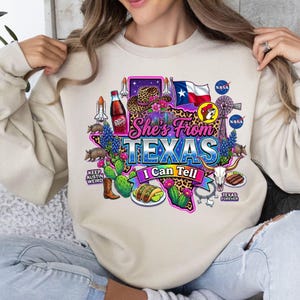 Pode incluir: Uma camisola bege de gola redonda com um gr&aacute;fico do estado do Texas com preenchimento de estampado de leopardo. O design inclui um chap&eacute;u de cowboy, uma garrafa de Dr Pepper, um vaiv&eacute;m espacial, a bandeira do Texas, tatus, tacos, um cr&acirc;nio de longhorn e o texto She's From TEXAS I Can Tell.