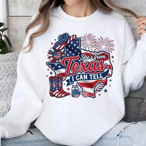 Pode incluir: Uma camisola branca de manga comprida com um gr&aacute;fico do estado do Texas com o padr&atilde;o da bandeira americana. O design inclui um chap&eacute;u de cowboy, botas, fogo de artif&iacute;cio, um frasco, &oacute;culos de sol e o texto She's from Texas I CAN TELL.