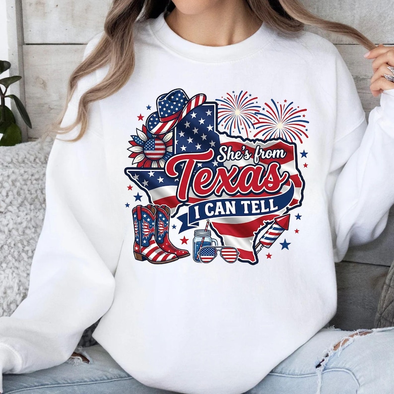 Pode incluir: Uma camisola branca de manga comprida com um gr&aacute;fico do estado do Texas com o padr&atilde;o da bandeira americana. O design inclui um chap&eacute;u de cowboy, botas, fogo de artif&iacute;cio, um frasco, &oacute;culos de sol e o texto She's from Texas I CAN TELL.