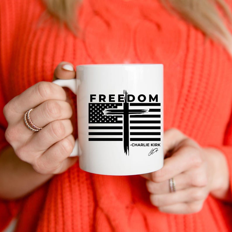 May include: White ceramic mug with the word "FREEDOM" in black above a design combining the American flag and a cross. The mug also has the text "-CHARLIE KIRK" and a signature. The mug is held by a person.