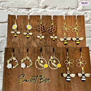 Puede incluir: Exhibidor de madera con varios pendientes dorados con diseños de abejas y flores. Algunos pendientes tienen diseños de margaritas y panales. El texto "Sweet Bee" es visible en la exhibición.