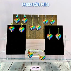 May include: A display of heart-shaped earrings and a bracelet in the colors of the rainbow pride flag. The earrings are on display stands with the text "Progressive Pride" at the top. The earrings have gold-colored hooks and posts.