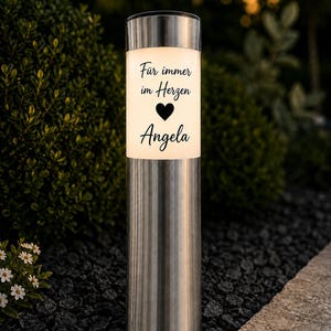 Könnte beinhalten: Eine zylindrische Solar-Gartenleuchte aus Edelstahl mit einem weiß beleuchteten Bereich, der den schwarzen Schriftzug "Für immer im Herzen", ein Herzsymbol und den Namen "Angela" in einer Schreibschrift zeigt.