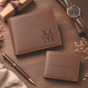Puede incluir: Una cartera plegable de cuero marrón grabada con el nombre Matthew y una letra M grande, junto a un tarjetero de cuero marrón más pequeño grabado con el texto Forever & Always, un bolígrafo marrón y un reloj de pulsera con correa de cuero marrón.