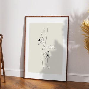 May include: A framed art print featuring a minimalist line drawing of two hands reaching towards each other. The hands have black nail polish. The print has the text "Love is two solitudes meeting at the edge". The frame is brown.