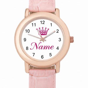 Pode incluir: Um relógio de pulso em ouro rosa com mostrador branco com uma coroa rosa e a palavra "Name" em escrita rosa. O relógio tem uma pulseira de couro rosa.