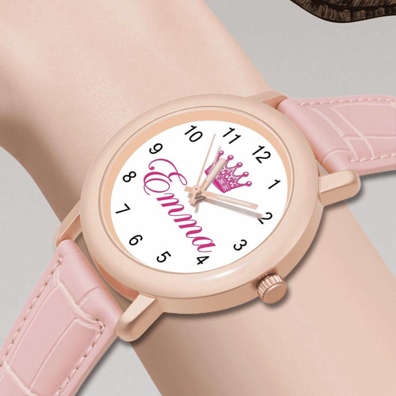 May include: A pink wristwatch with a white face featuring the name "Emma" in cursive and a pink crown graphic. The watch has a rose gold case and a pink leather strap.