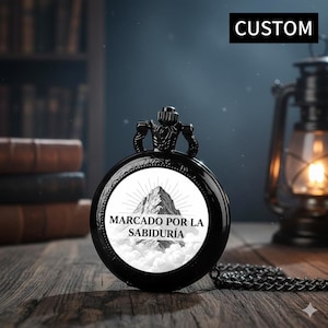 May include: Black pocket watch with a chain, featuring a mountain illustration and the text "MARCADO POR LA SABIDURÍA". The watch has a "CUSTOM" label and is set against a blurred background with books and a lantern.