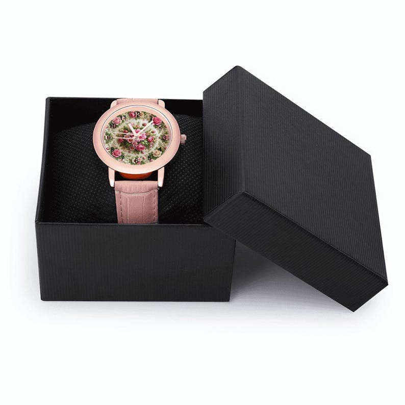 May include: A rose gold wristwatch with a floral face and a pink leather band, presented in an open black gift box. The watch face features a detailed design of pink and white flowers. The watch is displayed on a black cushion.