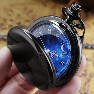 Puede incluir: Reloj de bolsillo negro con una esfera azul con temática de galaxia, con números romanos y un diseño de media luna. El reloj tiene una tapa con bisagras y una cadena. El reloj está abierto, revelando la esfera detallada.