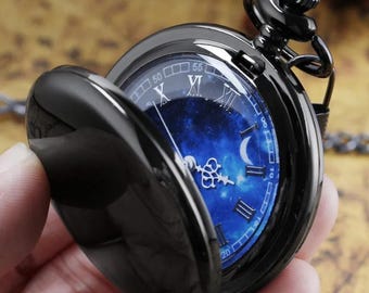 May include: Black pocket watch with a blue galaxy-themed face, featuring Roman numerals and a crescent moon design. The watch has a hinged cover and a chain. The watch is open, revealing the detailed face.