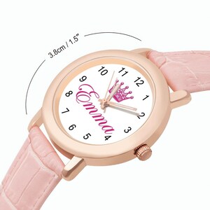 May include: A rose gold watch with a white face featuring the name "Emma" in pink script and a pink crown. The watch has a pink leather band and the face is 1.5 inches in diameter.