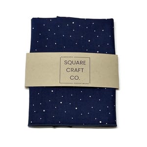 May include: A folded dark navy blue fabric square featuring a white polka dot pattern. A tan paper band wraps around the center, printed with the text "HANDMADE SQUARE CRAFT CO. POCKETWEAR" inside a rectangular border.