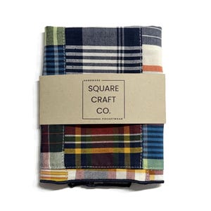 May include: A folded patchwork pocket square with a variety of plaid patterns in shades of blue, green, red, and yellow. A brown paper band with the text "SQUARE CRAFT CO. POCKETWEAR" wraps around the center of the pocket square.