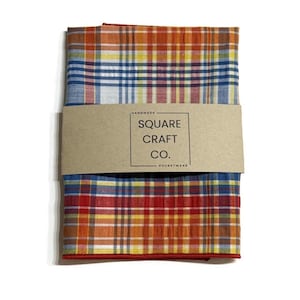 May include: A folded pocket square with a plaid pattern in shades of red, orange, yellow, and blue. A brown paper band wraps around the pocket square, with the text "SQUARE CRAFT CO. POCKETWEAR".