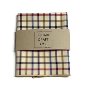 May include: A folded pocket square with a cream-colored background and a plaid pattern in burgundy, navy, gold, and brown. A brown paper band with the text "SQUARE CRAFT CO. POCKETWEAR" wraps around the center of the pocket square.
