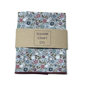 May include: A folded pocket square with a light blue background featuring a floral pattern in shades of pink, dark blue, and green. A tan paper band wraps around the center with the text "HANDMADE SQUARE CRAFT CO. POCKETWEAR".