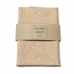 May include: A folded tan fabric square featuring a white polka dot pattern. A brown paper band wraps around the center, printed with the text: HANDMADE SQUARE CRAFT CO. POCKETWEAR inside a black rectangular border.