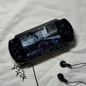 Puede incluir: Consola de juegos portátil Sony PSP negra que muestra un juego de carreras. La consola tiene un protector de pantalla y está conectada a auriculares negros. Un charm plateado con formas de estrella y hashtag cuelga de la consola.