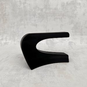 Könnte beinhalten: Eine schwarze, abstrakte Skulptur mit geschwungenem Design. Die Skulptur hat eine flache Basis und eine horizontale, rechteckige Oberseite, die durch einen geschwungenen Abschnitt verbunden sind. Das Objekt besteht aus einem glatten, reflektierenden Material.