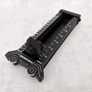 Könnte beinhalten: Ein schwarzes Präzisionsmesswerkzeug mit einem Schiebeelement, weißen Markierungen für Zoll und Zentimeter sowie dem Schriftzug CADEAUSWEDEN. Das Gerät verfügt über zwei silberfarbene, kreisförmige Akzente und einen zentralen Drehknopf.