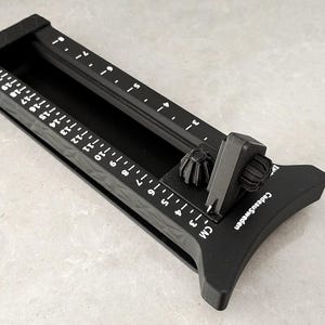Può includere: Un tagliacarte in plastica nera con un meccanismo di guida scorrevole. Il dispositivo presenta una scala metrica stampata che va da 0 a 20 cm sul lato. Il testo 'CadeauSweden' è stampato in bianco sulla base vicino al componente scorrevole.