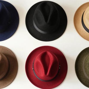 Op de afbeelding: Zes fedora hoeden in verschillende kleuren tegen een witte achtergrond. De hoeden zijn marineblauw, zwart, beige, bruin, rood en olijfgroen. Elke hoed heeft een band rond de kroon en de randen zijn iets omhoog gekruld.