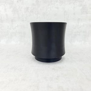 Peut inclure: Un vase en céramique noir avec un corps incurvé et une petite base circulaire. Le vase a une finition mate et un design simple et moderne. Le vase est vide et repose sur une surface blanche.