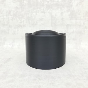 May include: A cylindrical, matte black object with a rounded top. The object has a smooth, uniform texture and appears to be made of a solid material. The design is simple and modern, with clean lines and a minimalist aesthetic.