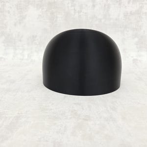 May include: A black, dome-shaped object with a cylindrical base. The object has a smooth, matte finish and is placed on a light grey surface. The shape suggests a potential lid or cover for a container.