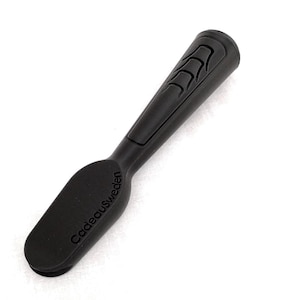 Könnte beinhalten: Ein mattschwarzes Handwerkzeug mit einem abgerundeten, flachen Ende und einem strukturierten, ergonomischen Griff. Das Wort CadeauSweden ist auf dem flachen Teil eingeprägt, und der Griff weist eine Reihe erhabener Rippen auf.