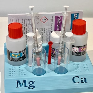 Op de afbeelding: Een lichtblauwe kunststof testkit-organizer met twee witte flessen met rode doppen gelabeld Mg-2 en Ca-2, twee doorzichtige plastic flesjes, twee injectiespuiten, een klein rood roerstaafje en twee instructiekaarten voor Calcium en Magnesium.