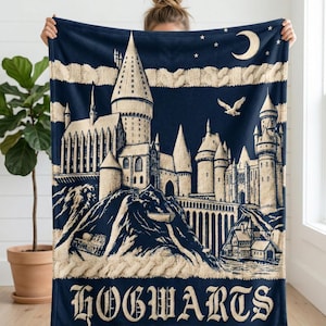 May include: Navy blue throw blanket featuring a detailed illustration of Hogwarts castle, with a crescent moon and stars. The word "HOGWARTS" is displayed in a decorative font. The blanket has a cable knit border.