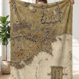 May include: A large, rectangular blanket featuring a detailed map of Middle Earth. The map is in shades of brown and beige, with intricate details of mountains, forests, and seas. The words "Middle Earth" are displayed in a decorative banner.