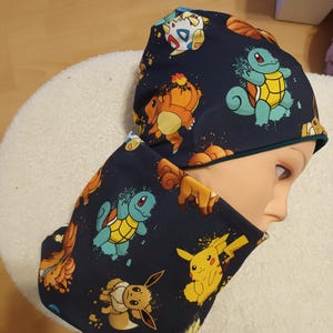 Könnte beinhalten: Ein dunkelblaues Beanie und ein passender Schlauchschal mit einem bunten Allover-Print verschiedener animierter Charaktere wie Pikachu, Schiggy, Evoli und Togepi. Der Stoff hat eine glatte Textur und das Set wird auf einem Puppenkopf präsentiert.